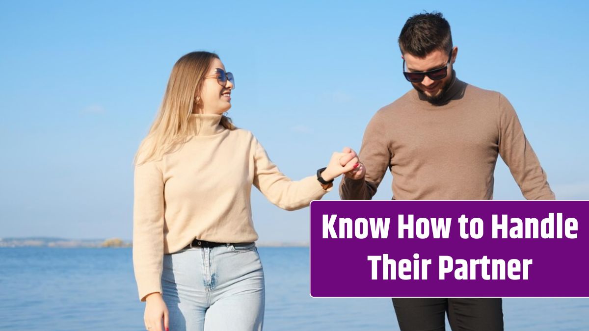 Young couple in jeans and sweaters holding hands and walking along the beach in sunny weather.